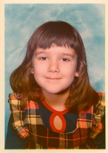 Retrato de una niña con cabello castaño y flequillo, vestida con un diseño a cuadros rojo, amarillo y negro sobre blusa azul con cuello rojo. Fondo azul claro