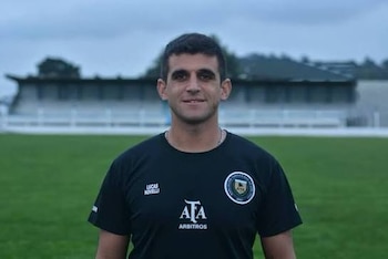 Un hombre con camiseta negra de la AFA posa frente a un campo de fútbol con gradas al fondo bajo un cielo nublado