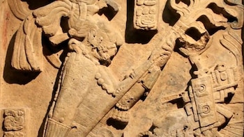 Primer plano de un relieve en piedra caliza con dos figuras antropomorfas de perfil, vestimenta detallada, tocados y ornamentos