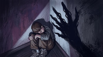 Un niño sentado en el suelo de un rincón oscuro abraza sus piernas cerca de una sombra de dedos largos proyectada en la pared.