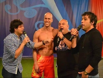 Cuatro hombres de pie en un escenario. Uno lleva camiseta a cuadros, uno está sin camiseta, uno lleva camiseta negra con logo, y otro lleva camiseta negra. Dos sostienen micrófonos