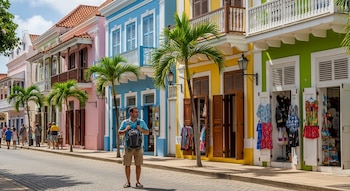 Hombre con mochila de pie en calle empedrada rodeado de edificios de colores pastel con balcones, ventanas y puertas de madera, y palmeras.