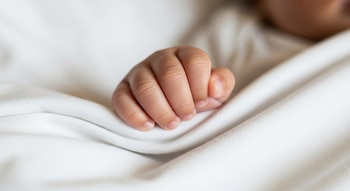 Primer plano de mano de bebé con dedos cerrados y piel clara sobre una sábana blanca con pliegues. Parte de la cabeza del bebé es visible al fondo.