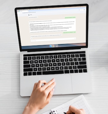 Vista superior de una persona usando un portátil plateado con la pantalla mostrando un chat de IA y una mano escribiendo en un cuaderno con espiral