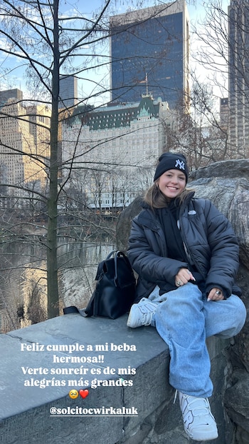 Una joven sonríe, sentada en una pared de piedra, viste gorro negro con logo NY, chaqueta acolchada, jeans, zapatillas. Fondo con árboles y edificios