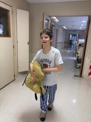 Un niño con camiseta gris y pantalón a cuadros camina con un muñeco de peluche amarillo por el pasillo de un centro hospitalario