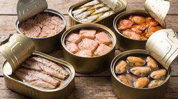 Seis latas metálicas abiertas que contienen atún, salmón, sardinas y mejillones sobre una tabla de madera.