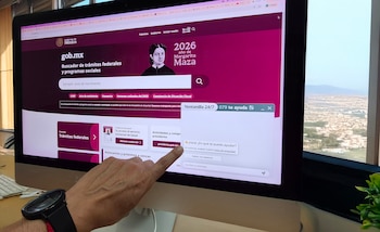 Pantalla de computadora muestra el portal gob.mx del Gobierno de México, con una mano señalando. Se ve un chat de ayuda y un paisaje urbano a través de una ventana