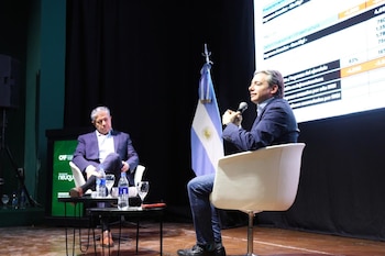 Dos hombres sentados en un escenario junto a la bandera de Argentina, una mesa con agua y una pantalla con datos proyectados