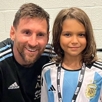 Lionel Messi con barba sonríe junto a un niño con cabello largo y camisa celeste y blanca de Argentina, ambos mirando a cámara