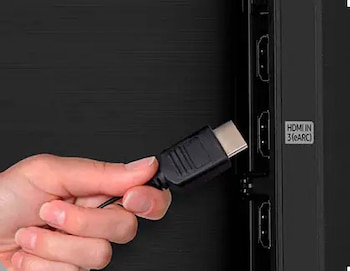 Una mano sostiene un cable HDMI, acercándolo a los puertos de entrada de un televisor negro. Se observa el texto 'HDMI IN 3 (eARC)'