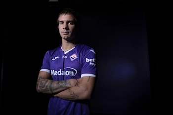 Hombre joven con camiseta de fútbol morada de la Fiorentina, con brazos cruzados y tatuajes visibles, sobre un fondo oscuro