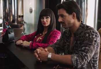 Mujer de cabello oscuro con flequillo y chaqueta fucsia, hombre de bigote con camisa estampada, sentados en una mesa oscura, con bolso negro y pañuelo
