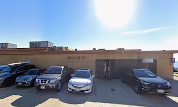Fachada de un edificio con el letrero NOBU y varios automóviles estacionados al frente bajo un cielo despejado