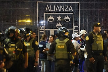 Agentes de policía con equipo táctico y personas se encuentran frente a una pared de azulejos oscuros con el logo blanco del Club Alianza Lima