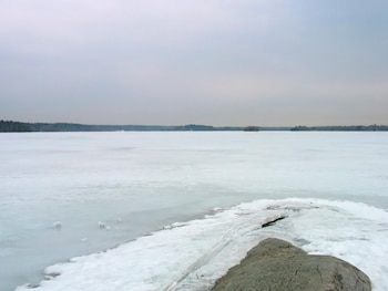 Imagen panorámica de un lago congelado, con una gran roca gris y nieve en primer plano, y un bosque oscuro al fondo bajo un cielo gris