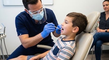 Odontólogo con mascarilla, protector facial y guantes azules examina la boca de un niño sentado en un sillón dental. Una mujer sentada en el fondo.
