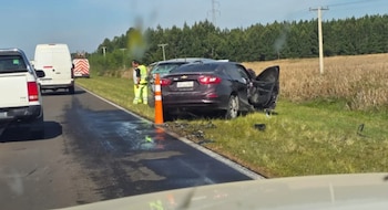 Un automóvil oscuro dañado sobre el césped al costado de una carretera, junto a un cono de tránsito naranja y vehículos en la vía
