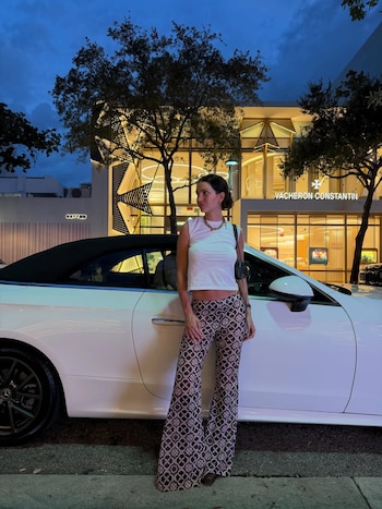 Zaira Nara de pie junto a un coche descapotable blanco en una calle de noche. Viste un top blanco y pantalones estampados, con una tienda iluminada al fondo