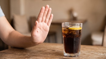 Una mano abierta en gesto de detención frente a un vaso de gaseosa con hielo y una rodaja de limón sobre una mesa de madera.