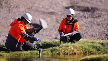 Un hombre y una mujer con cascos blancos y chaquetas de seguridad miden la calidad del agua con un sensor en un arroyo