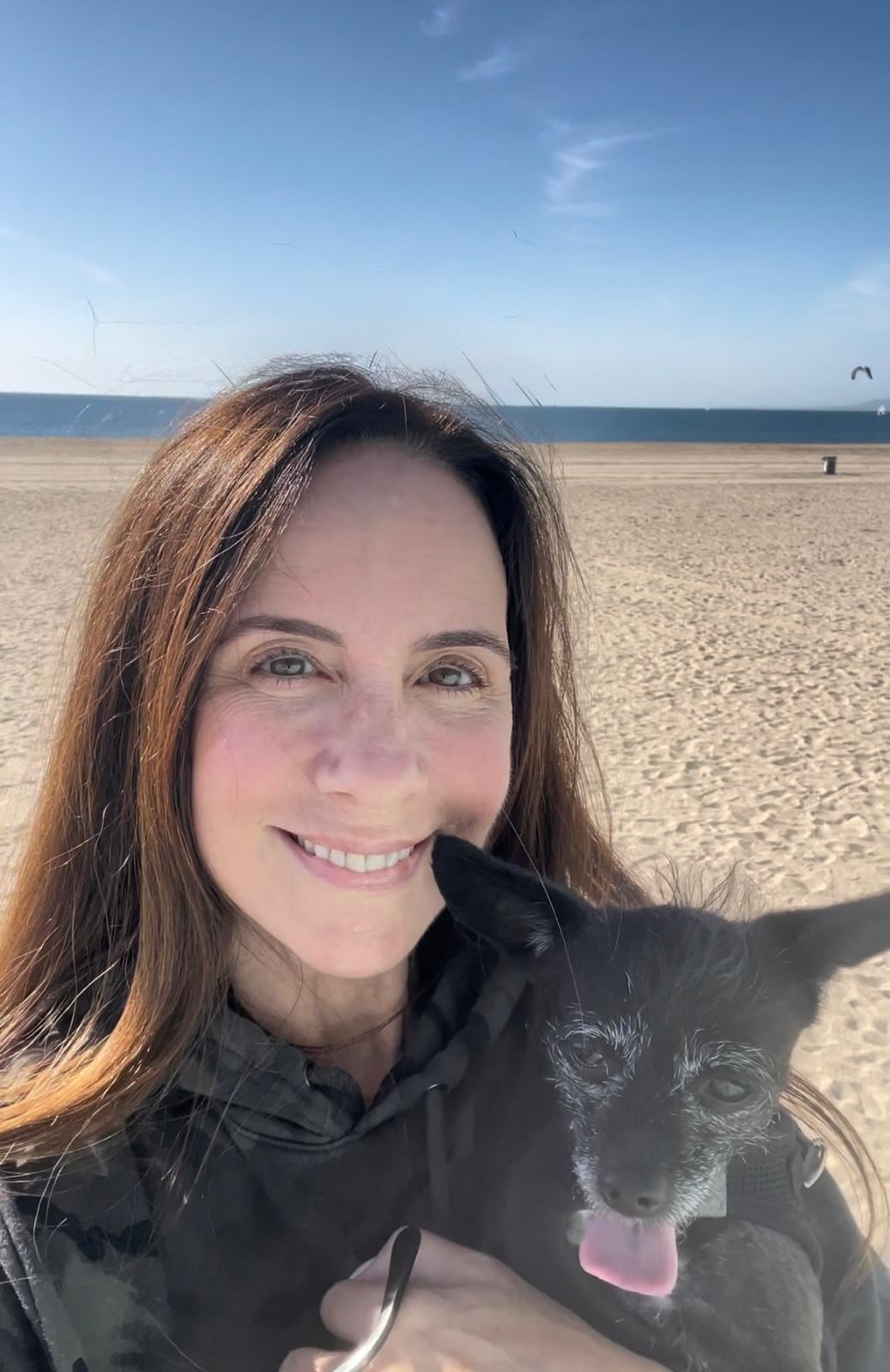 Noelle Balfour sonríe mientras sostiene a un perro pequeño en una playa abierta frente al mar.