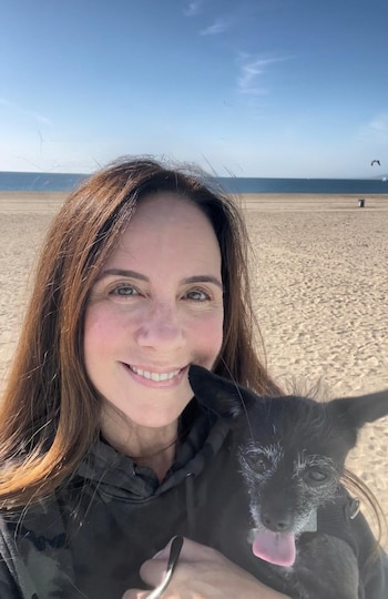 Una mujer de cabello castaño sonríe a la cámara mientras sostiene un perro negro pequeño en una playa de arena