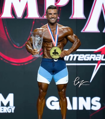 Fisicoculturista sonriente en el escenario sostiene un trofeo de cristal y muestra una medalla dorada colgada al cuello