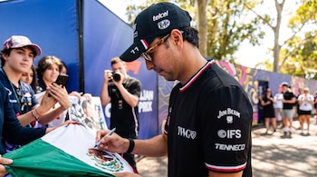 Un hombre con gorra y camiseta oscura firma una bandera de México. Aficionados sostienen la bandera y usan sus teléfonos. Un fotógrafo detrás.
