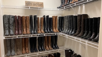 Pares de botas altas alineados en estanterías de alambre blanco en tonos negros, marrones y grises, con un cartel que dice Mis botas.