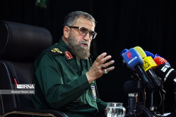 Hombre con barba y gafas en uniforme militar verde habla frente a cinco micrófonos de prensa. Hay un vaso de agua