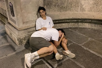 Dos personas sentadas en el suelo de baldosas junto a un pilar de piedra. Una persona apoya su cabeza en el regazo de la otra, que observa hacia un lado