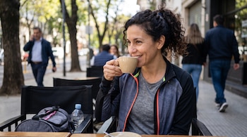 Mujer con cabello rizado en coleta, chaqueta deportiva negra y gris, bebiendo de una taza beige en una mesa de madera. Una mochila gris y una botella de agua están al lado.