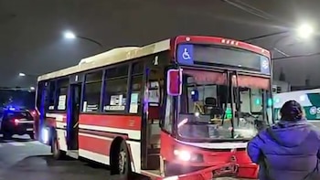 Vista nocturna de un colectivo rojo y blanco con daños frontales. Un patrullero con luces azules y una ambulancia están cerca. Una persona de espaldas observa