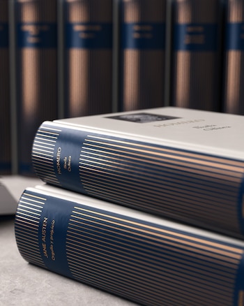 Dos libros de tapa dura con lomos azul marino y detalles dorados están apilados; se leen "Homero" y "Jane Austen"; hay una fila de libros similares desenfocados detrás