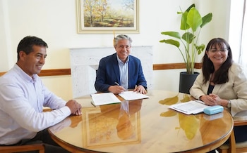 Tres personas sonrientes sentadas alrededor de una mesa redonda de madera con papeles, frente a una chimenea y una planta