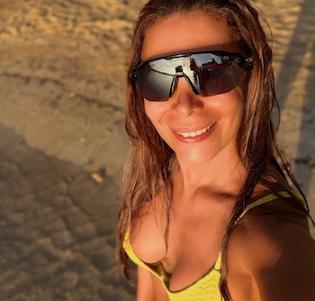 Una mujer con gafas de sol oscuras y traje de baño amarillo sonríe en una playa de arena durante el atardecer