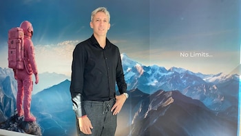 Un hombre de cabello gris con camisa negra y pantalones grises de pie frente a un mural azul con montañas, un astronauta rosa y texto 'No Limits...'