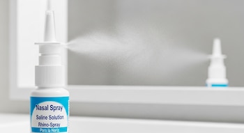 Primer plano de un frasco blanco de aerosol nasal de solución salina que rocía una fina niebla hacia la derecha, con otro frasco similar desenfocado al fondo.