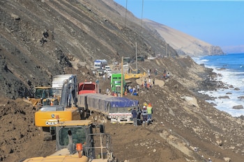 Maquinaria pesada, camiones y personas en un tramo de carretera costera cubierto por tierra y piedras junto al mar