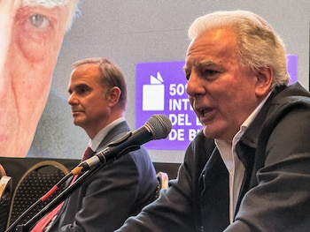 Un hombre de cabello canoso habla frente a un micrófono, con un hombre de cabello oscuro al fondo y una pantalla detrás con un ojo y texto parcialmente visible