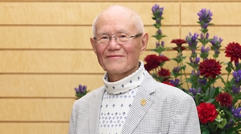 Un hombre de edad avanzada con gafas redondas y saco a cuadros posa frente a una pared de madera con flores rojas y moradas.