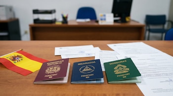 Tres pasaportes abiertos (granate, azul, verde) con escudos, una bandera de España y documentos dispersos sobre una mesa de madera clara en una oficina.