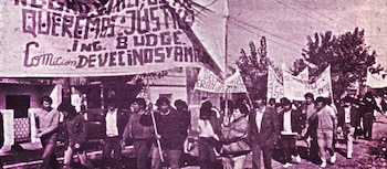 Una fotografía antigua con tono púrpura muestra a un grupo de personas marchando, llevando un gran cartel blanco con texto oscuro y otras pancartas