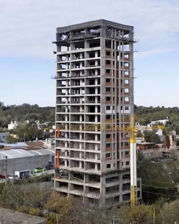 Vista aérea de un edificio de hormigón de muchos pisos sin terminar, con andamios visibles en la parte superior, rodeado de árboles y edificios bajos