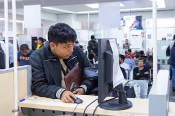 Un hombre joven sentado en un escritorio usa un ordenador personal con un monitor y un ratón. Al fondo, otras personas esperan en una sala