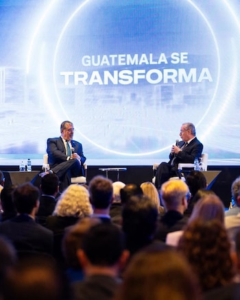 Dos hombres sentados en sillas, micrófonos, pantalla luminosa con texto "GUATEMALA SE TRANSFORMA", mesas con botellas de agua, audiencia borrosa
