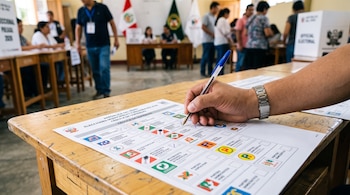Una mano sostiene un bolígrafo azul y marca una cédula de sufragio electoral colocada sobre una mesa de madera.