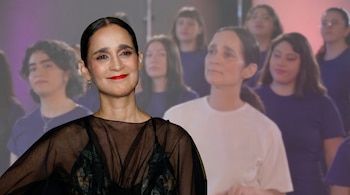 Una mujer de cabello recogido y blusa transparente negra sonríe mirando al frente, con labios rojos, mientras un grupo de personas con camisetas moradas y claras aparece difuminado al fondo.