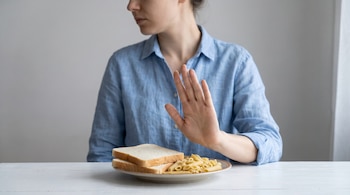 Mujer sentada con camisa azul, mesa blanca, plato con dos rebanadas de pan blanco y pasta penne, mano levantada en señal de rechazo, fondo gris.
